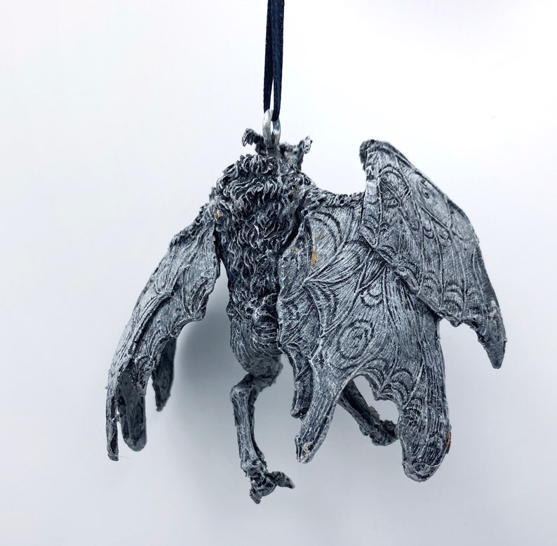 Puede incluir: Un adorno gris, detallado, con forma de murci&eacute;lago con un cord&oacute;n negro que cuelga de la parte superior. El murci&eacute;lago tiene un aspecto texturizado, casi esquel&eacute;tico, con intrincados dise&ntilde;os de alas.