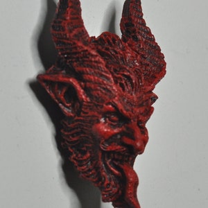 May include: A red, detailed, ceramic sculpture of a devil's head with horns and a snarling expression.