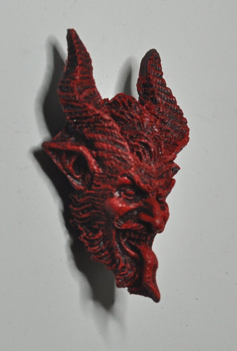 May include: A red, detailed, ceramic sculpture of a devil's head with horns and a snarling expression.
