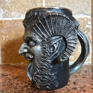 May include: A silver and black ceramic mug with a detailed sculpted face of a mythical creature with horns and a beard. The mug has a handle and a silver rim.