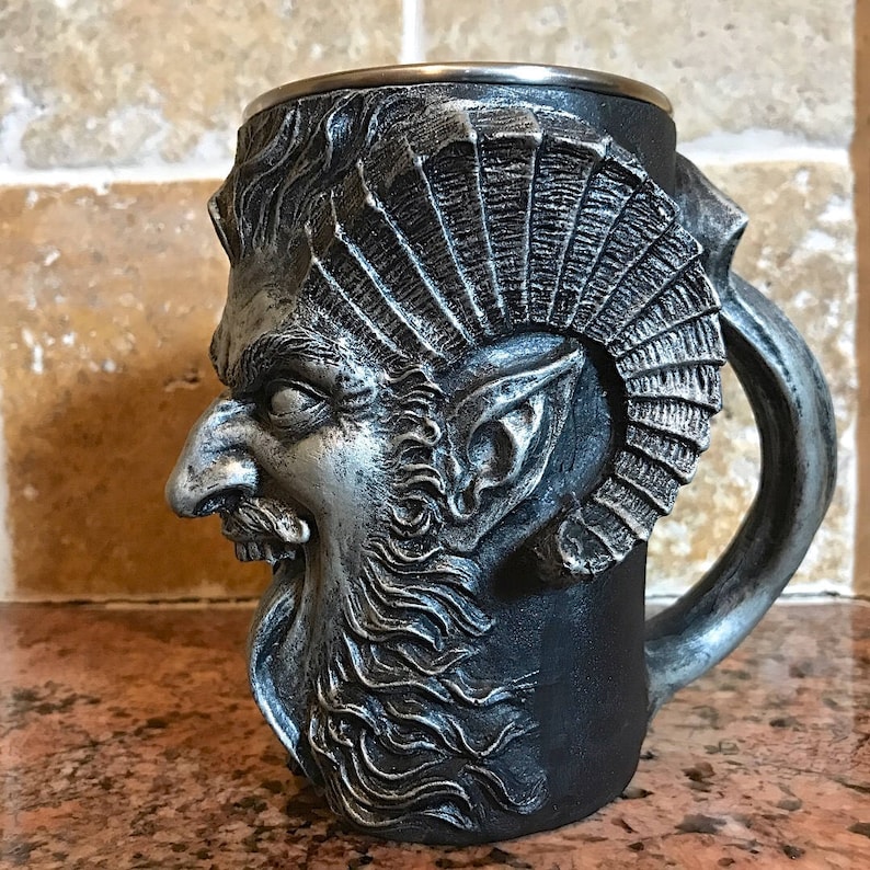 May include: A silver and black ceramic mug with a detailed sculpted face of a mythical creature with horns and a beard. The mug has a handle and a silver rim.