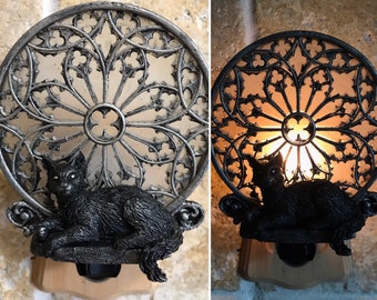 May include: A black cat figurine sits inside a decorative metal nightlight shaped like a gothic window. The nightlight is plugged in and lit, casting a warm glow.