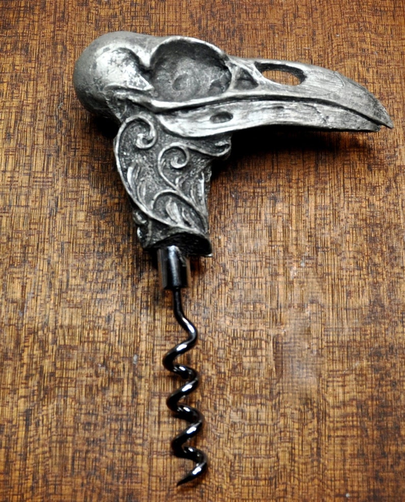 May include: A silver metal corkscrew shaped like a bird skull with intricate floral designs. The corkscrew has a black metal spiral and is on a brown wooden surface.