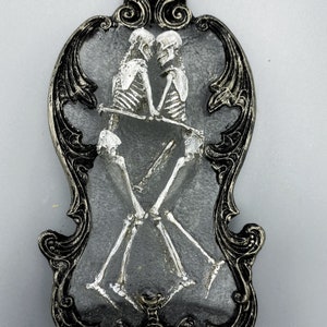 May include: A silver-painted skeletal couple embracing, framed in a black ornate frame with silver accents.