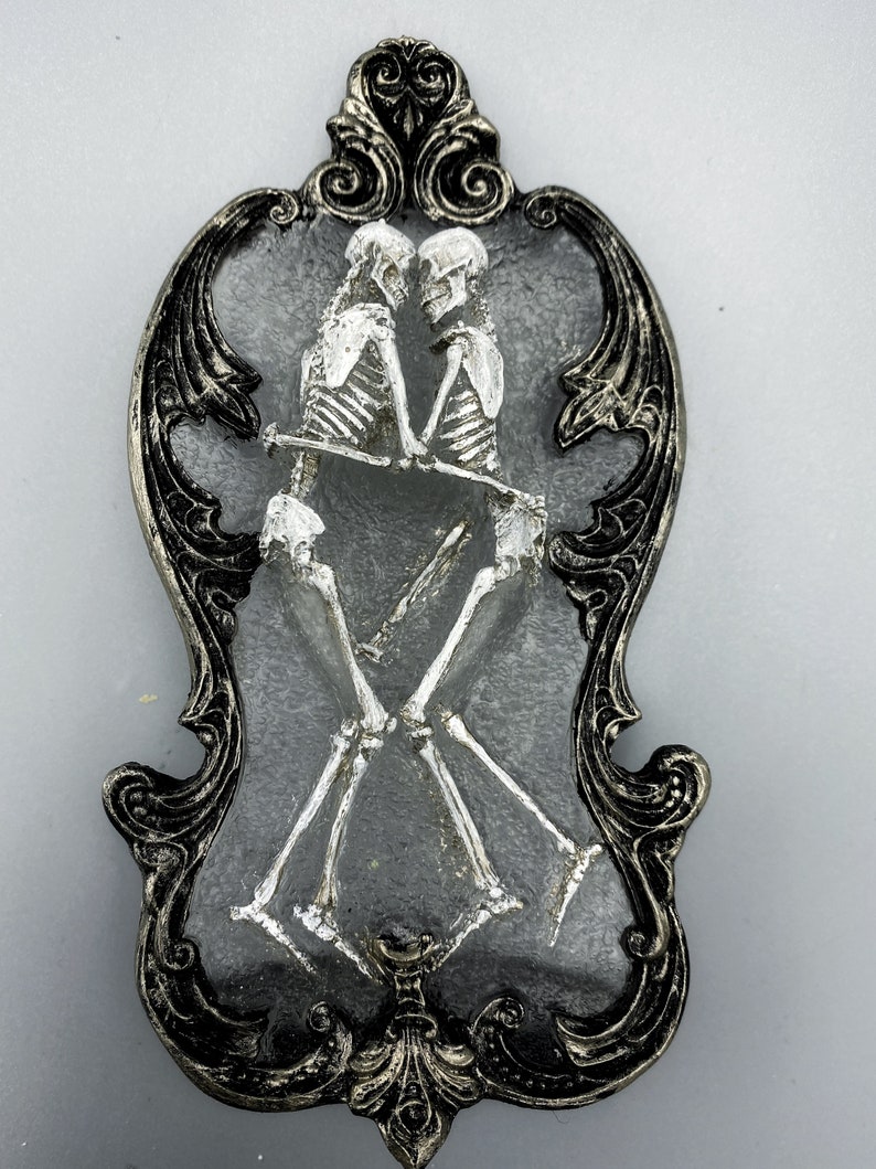 May include: A silver-painted skeletal couple embracing, framed in a black ornate frame with silver accents.