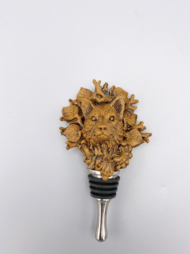 Op de afbeelding: Een bruine en gouden flessenstop met een gedetailleerd ontwerp van een wolfskop. De wolf is omringd door bladeren en takken. De stop heeft een zilveren metalen basis.