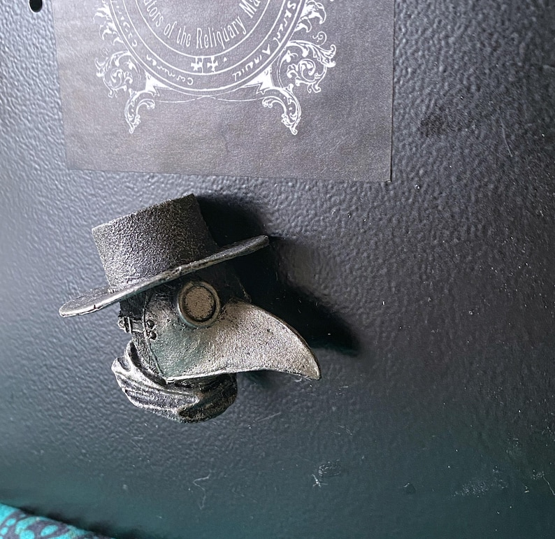 Pu&ograve; includere: Una maschera di medico della peste color argento con un cappello nero e una lunga sciarpa fluente. La maschera ha un becco grande e rotondo e un piccolo foro rotondo per gli occhi.
