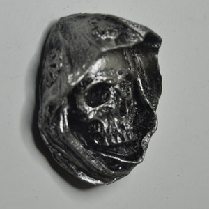 May include: A silver-colored skull sculpture with a hooded cloak. The skull has detailed features and is set against a white background.