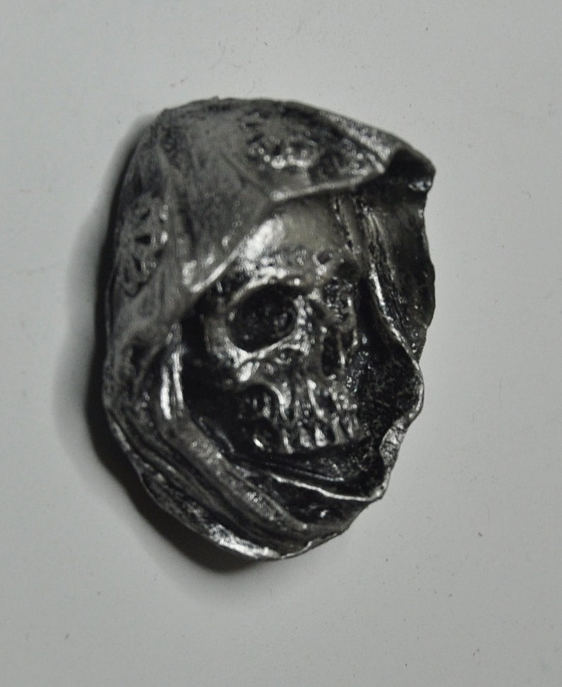 May include: A silver-colored skull sculpture with a hooded cloak. The skull has detailed features and is set against a white background.