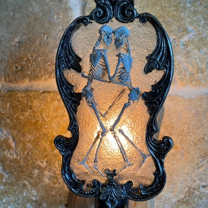 May include: A black and white nightlight with a decorative frame and a frosted glass panel featuring a couple of skeletons holding hands. The skeletons are in a romantic pose, with the male skeleton holding the female skeleton's hand. The nightlight is lit, casting a warm glow.