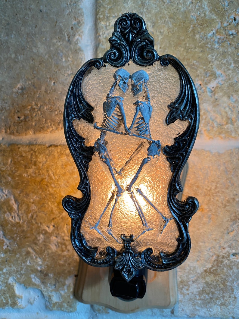 May include: A black and white nightlight with a decorative frame and a frosted glass panel featuring a couple of skeletons holding hands. The skeletons are in a romantic pose, with the male skeleton holding the female skeleton's hand. The nightlight is lit, casting a warm glow.