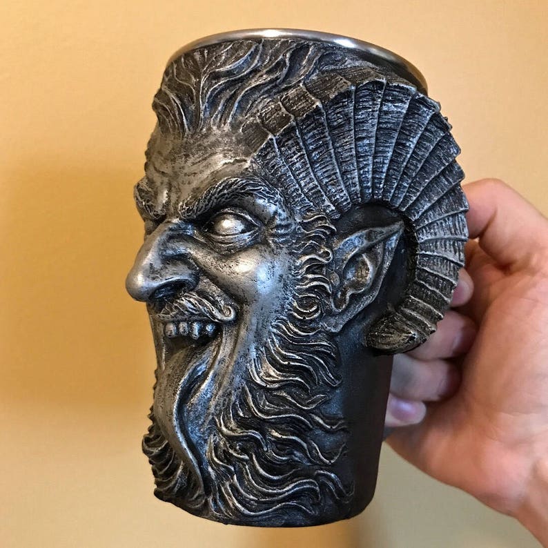 May include: A silver-colored metal mug shaped like a demon's head with horns, a beard, and a long tongue sticking out. The mug is made of a dark material and has a textured surface.