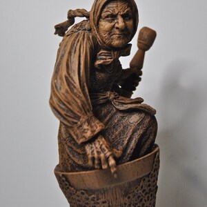 May include: A wooden sculpture of a woman with a weathered face, wearing a long brown robe and holding a wooden mallet. The sculpture is sitting on a wooden base with a detailed design.