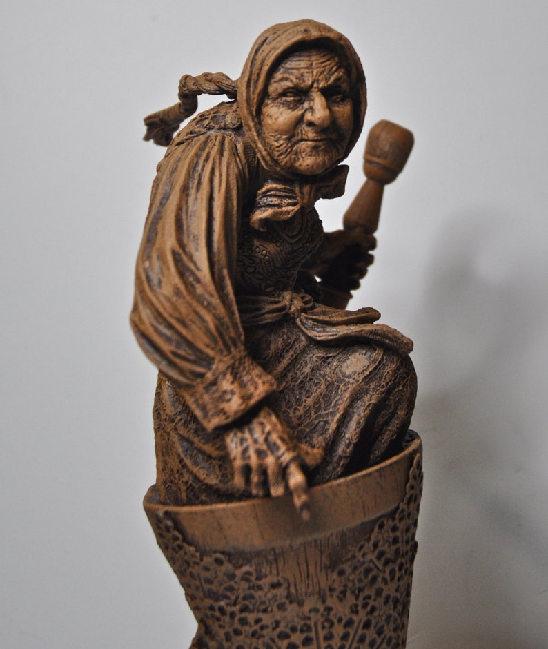 May include: A wooden sculpture of a woman with a weathered face, wearing a long brown robe and holding a wooden mallet. The sculpture is sitting on a wooden base with a detailed design.