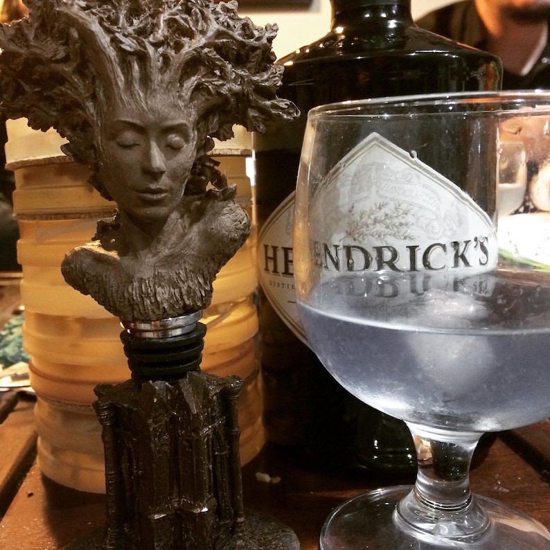 May include: A glass bottle of Hendricks Gin with a label that reads "Hendricks Gin" and a decorative bottle stopper featuring a sculpted woman's head with a tree-like hair style.