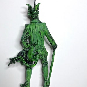 May include: Green figurine of a man with horns and a tail, wearing a long coat and holding a cane. The figurine is made of plastic and has a distressed paint finish.