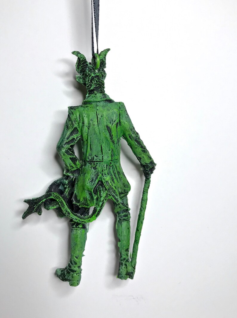 May include: Green figurine of a man with horns and a tail, wearing a long coat and holding a cane. The figurine is made of plastic and has a distressed paint finish.