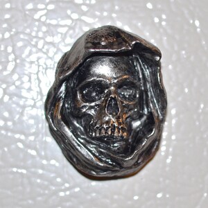 May include: A silver skull wearing a hooded cloak. The skull has a detailed face with visible eye sockets, a nose, and teeth.