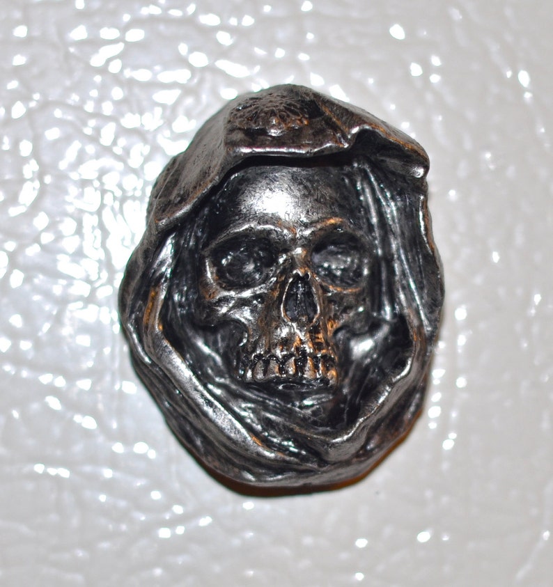 May include: A silver skull wearing a hooded cloak. The skull has a detailed face with visible eye sockets, a nose, and teeth.