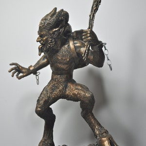 May include: A bronze statue of a goat-like creature with horns, a chain, and a stick in its hand. The creature is standing on a rocky base.