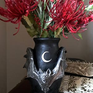 May include: Black ceramic vase with a bat and crescent moon design. The vase is filled with red and white artificial flowers.