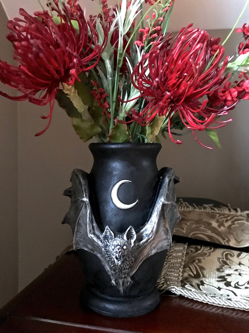 May include: Black ceramic vase with a bat and crescent moon design. The vase is filled with red and white artificial flowers.