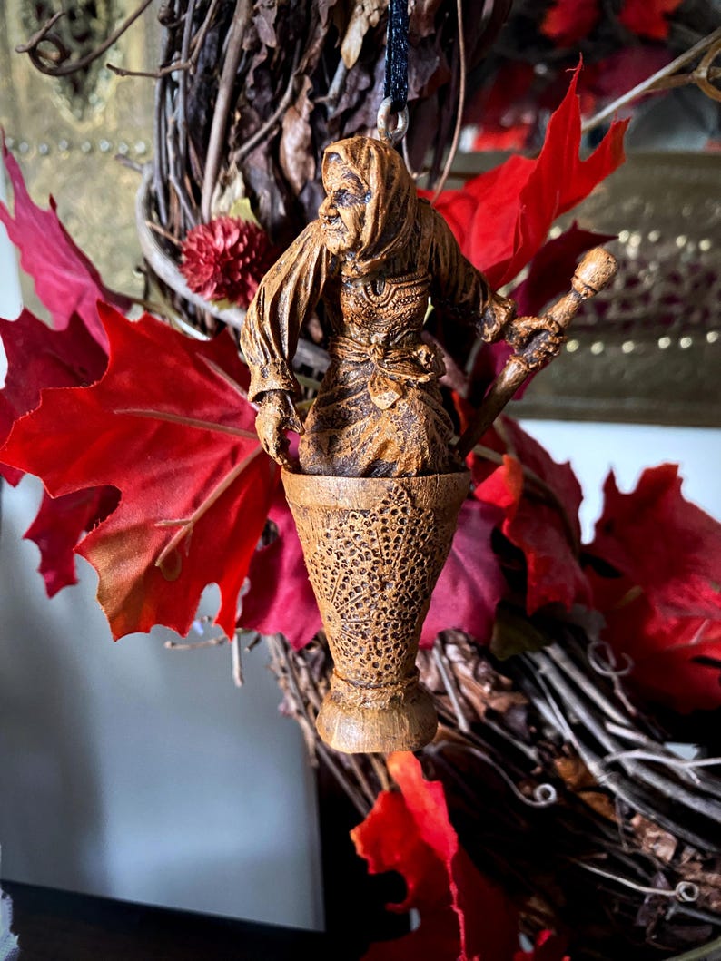 May include: A wooden ornament of a witch holding a staff, standing in a carved wooden cauldron. The ornament is brown and has a rustic look.