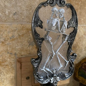 May include: A silver and black nightlight with a decorative frame and a clear glass insert featuring two skeletons embracing.