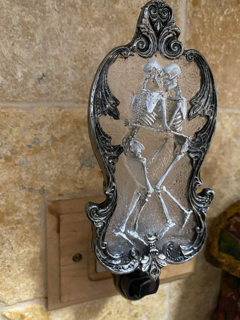 May include: A silver and black nightlight with a decorative frame and a clear glass insert featuring two skeletons embracing.