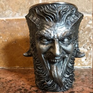 May include: A silver metal drinking cup shaped like a demon's head with horns, a beard, and a tongue sticking out.