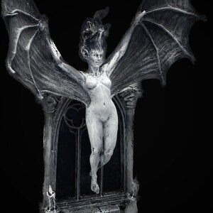 Pode incluir: Uma figura branca esculpida de uma mulher com asas, de p&eacute; em uma porta com arco pontiagudo. A figura est&aacute; nua e tem cabelo comprido e solto. A porta &eacute; decorada com uma janela de vitral e dois casti&ccedil;ais.