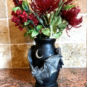May include: Black ceramic vase with a bat and crescent moon design. The vase is decorated with a gray bat with outstretched wings and a white crescent moon. The vase is filled with red and green artificial flowers.