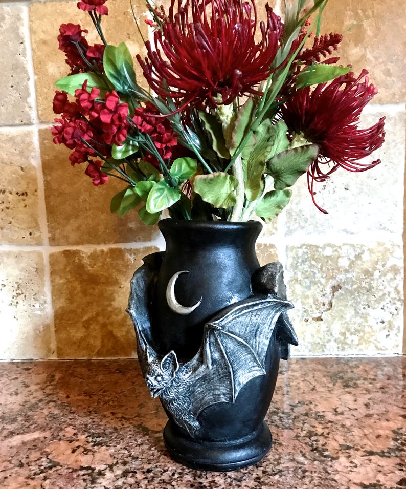 May include: Black ceramic vase with a bat and crescent moon design. The vase is decorated with a gray bat with outstretched wings and a white crescent moon. The vase is filled with red and green artificial flowers.