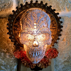 May include: A white ceramic skull with gold detailing and a floral design. The skull is illuminated from within, casting a warm glow. The skull is surrounded by red flowers and is mounted on a wooden base.