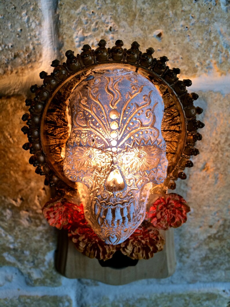 May include: A white ceramic skull with gold detailing and a floral design. The skull is illuminated from within, casting a warm glow. The skull is surrounded by red flowers and is mounted on a wooden base.