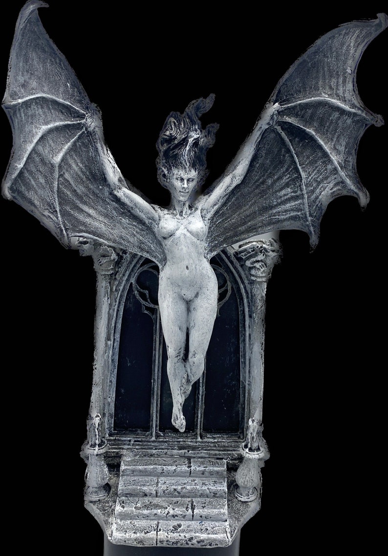 Pode incluir: Uma escultura de uma figura feminina alada branca com um arco g&oacute;tico atr&aacute;s. A escultura &eacute; feita de um material texturizado e tem um aspecto desgastado. A figura est&aacute; em p&eacute; em um conjunto de degraus com dois casti&ccedil;ais de cada lado.