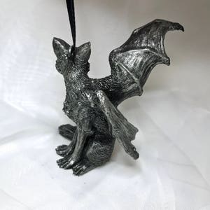 May include: A silver-colored figurine of a gargoyle with bat wings. The gargoyle is sitting with its wings spread out behind it.