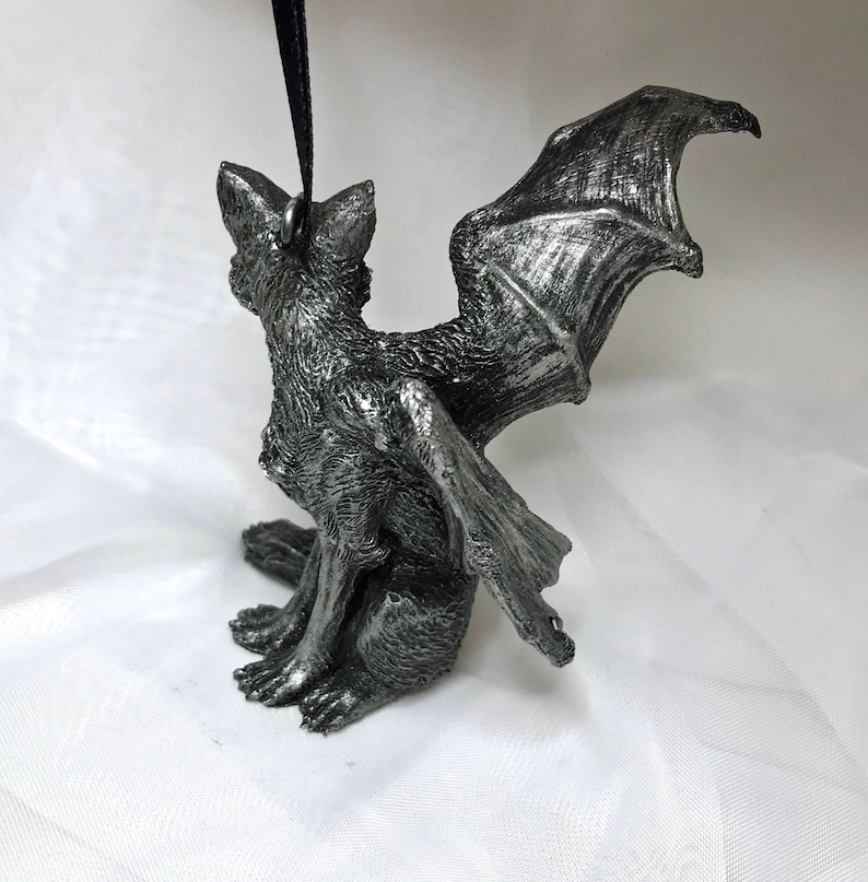 May include: A silver-colored figurine of a gargoyle with bat wings. The gargoyle is sitting with its wings spread out behind it.