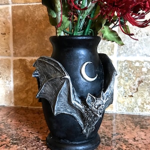 May include: Black ceramic vase with a bat and crescent moon design. The vase is decorated with a silver-colored bat and moon. The vase is filled with red flowers.