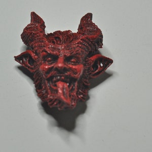 May include: A red, detailed, ceramic figurine of a devil with horns, a beard, and a protruding tongue.