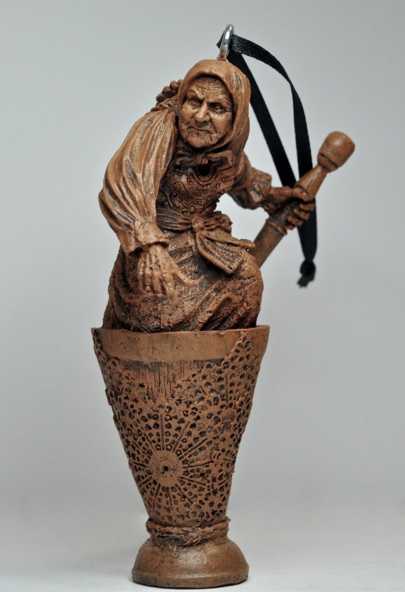 May include: A brown ceramic ornament of a woman holding a staff, standing in a brown ceramic cup with a decorative pattern. The ornament has a black ribbon for hanging.