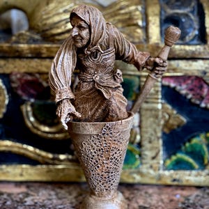 May include: A brown wooden figurine of a person wearing a hooded cloak and holding a staff. The figurine is standing in a brown wooden cup with a decorative design.