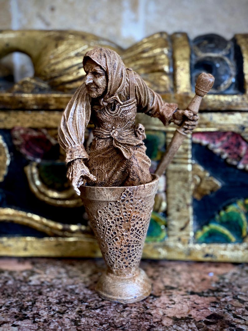 May include: A brown wooden figurine of a person wearing a hooded cloak and holding a staff. The figurine is standing in a brown wooden cup with a decorative design.