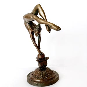 May include: A bronze sculpture of a woman performing a contortionist pose, balancing on a ball atop a decorative base.
