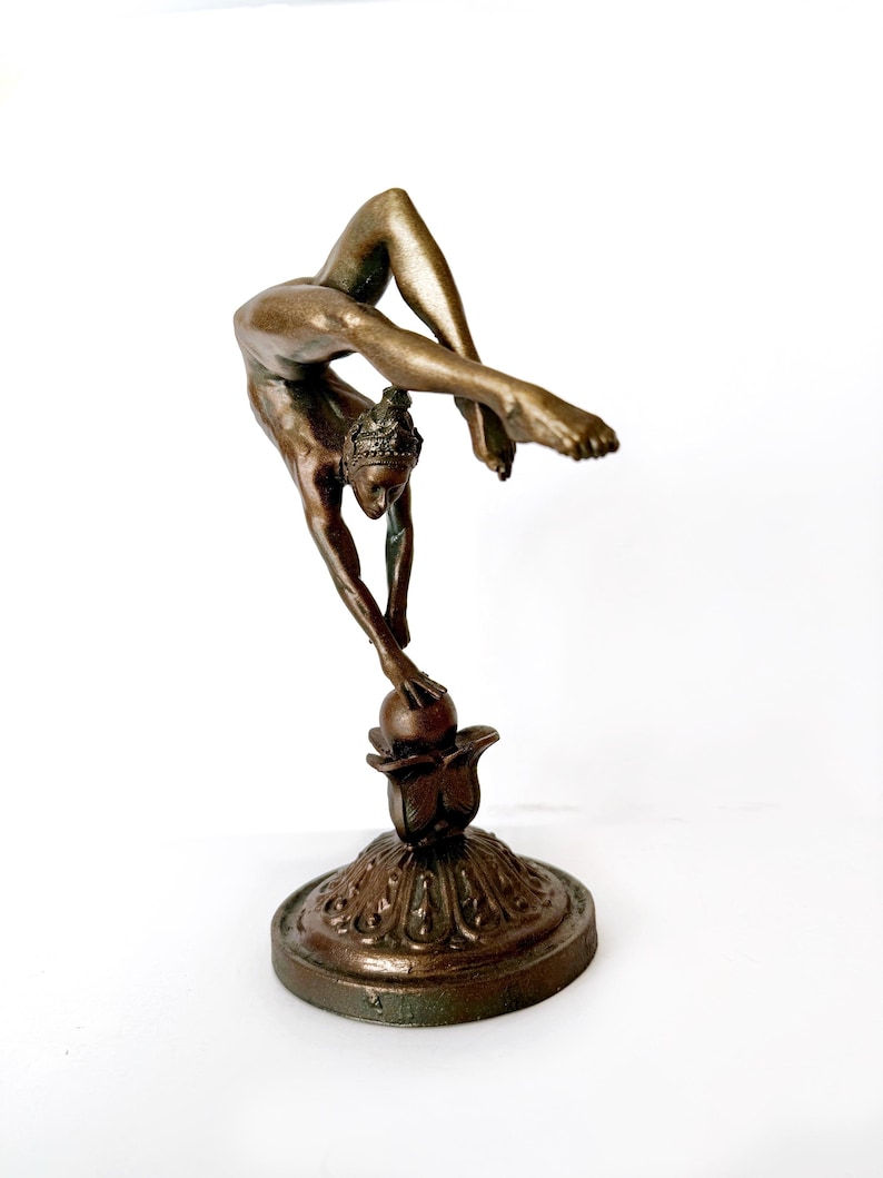 May include: A bronze sculpture of a woman performing a contortionist pose, balancing on a ball atop a decorative base.