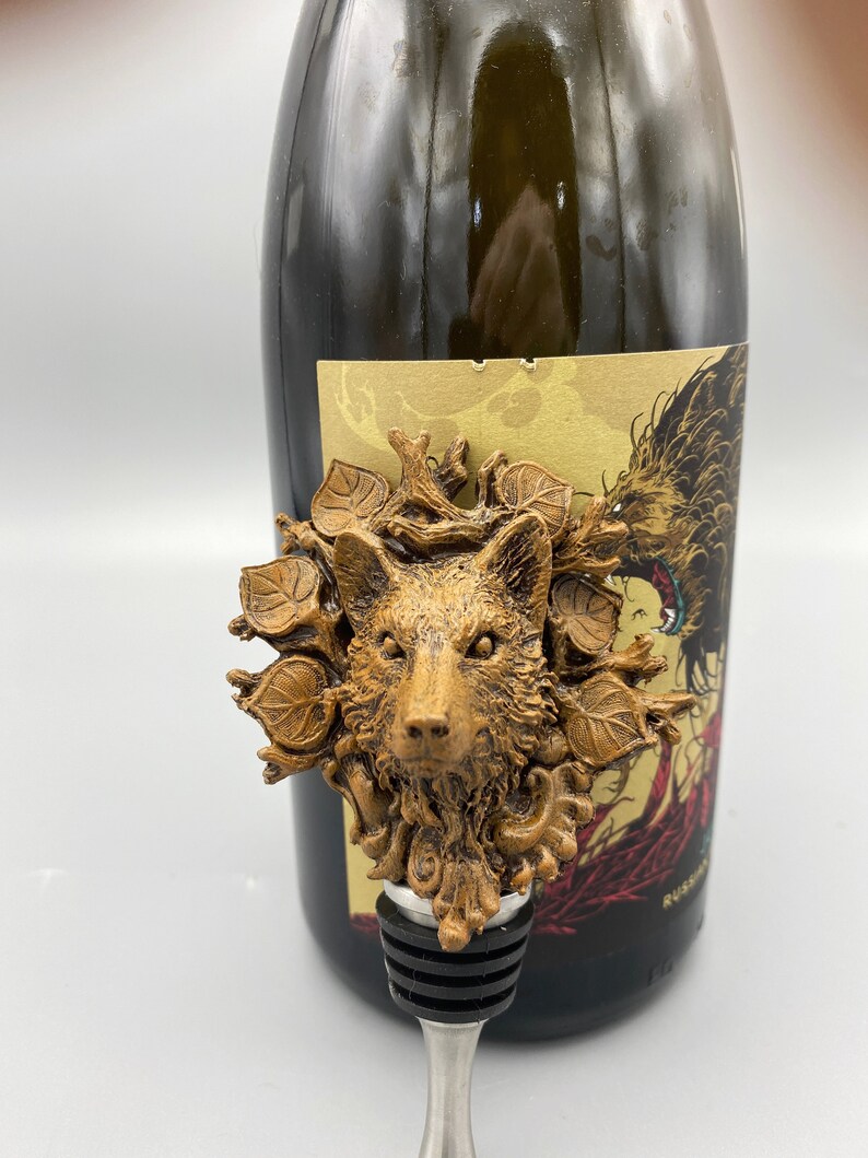 Op de afbeelding: Een gouden flesstopper met een wolvenkop omringd door bladeren. De stopper is gemaakt van metaal en heeft een zwarte rubberen afdichting. De wolvenkop is gedetailleerd en realistisch.