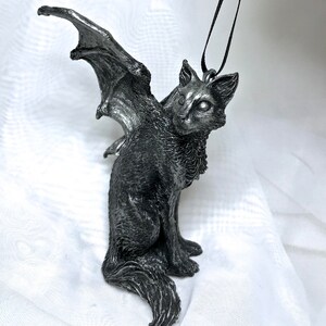 May include: A silver-colored winged cat ornament with a black cord for hanging. The cat is sitting with its wings spread out behind it.