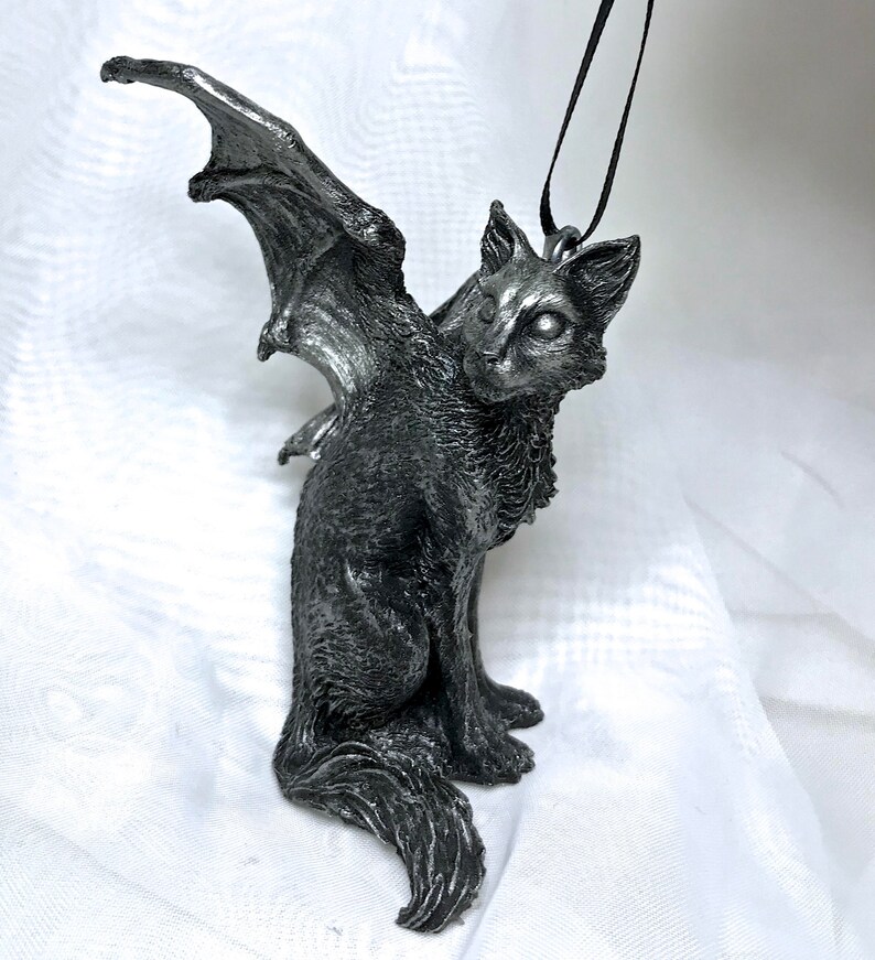 May include: A silver-colored winged cat ornament with a black cord for hanging. The cat is sitting with its wings spread out behind it.