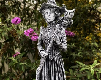 Puede incluir: Una figura de bruja gris y blanca con un sombrero negro y un búho posado en su hombro. La figura sostiene un bastón en su mano derecha.
