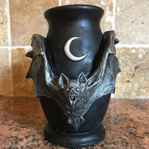 May include: Black ceramic vase with a bat and crescent moon design. The bat is silver and has a detailed design. The vase has a wide mouth and a rounded base.